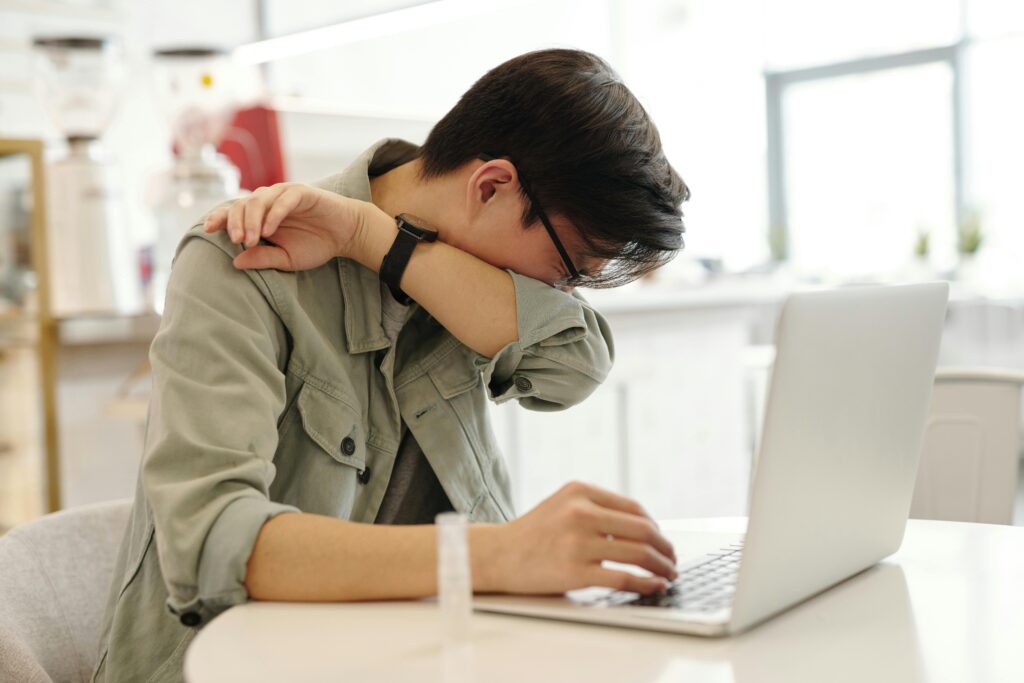 pexels-photo-4031631-4031631 A man sneezes into his elbow while using a laptop, depicting common cold symptoms in a modern workspace.