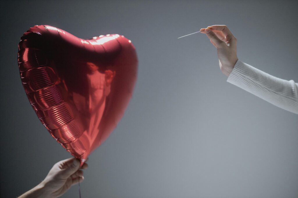 pexels-photo-7670313-7670313 Close-up of a red heart-shaped balloon being popped by a needle, evoking themes of love and fragility.